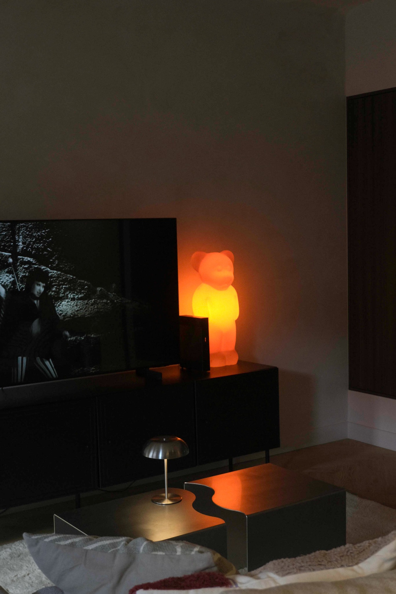 Nowoczesny salon z czarnymi meblami, telewizorem i lampą w kształcie misia, oświetlającą wnętrze ciepłym, pomarańczowym światłem. Metalowa lampka na stoliku kawowym.