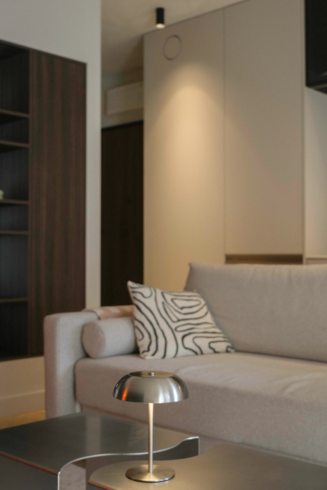 Minimalistyczny salon: beżowa sofa z wzorzystą poduszką, metalowa lampka na stoliku, ciemne regały w tle. Stonowana kolorystyka, nowoczesny design.