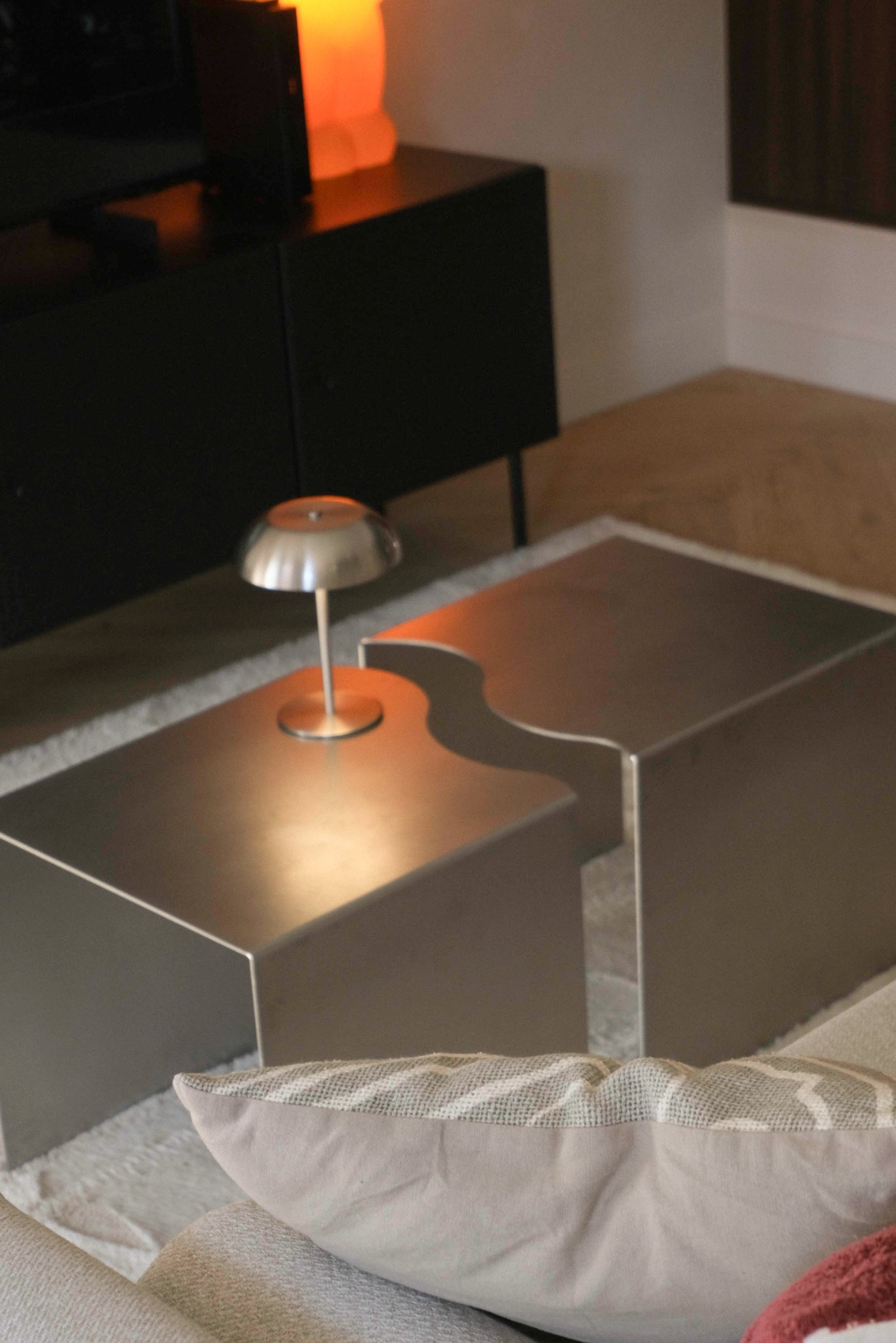 Nowoczesny stolik kawowy z metalu, o nieregularnym kształcie, z lampką. W tle czarna komoda i ciepłe światło lampy solnej. Poduszka dekoracyjna na sofie.