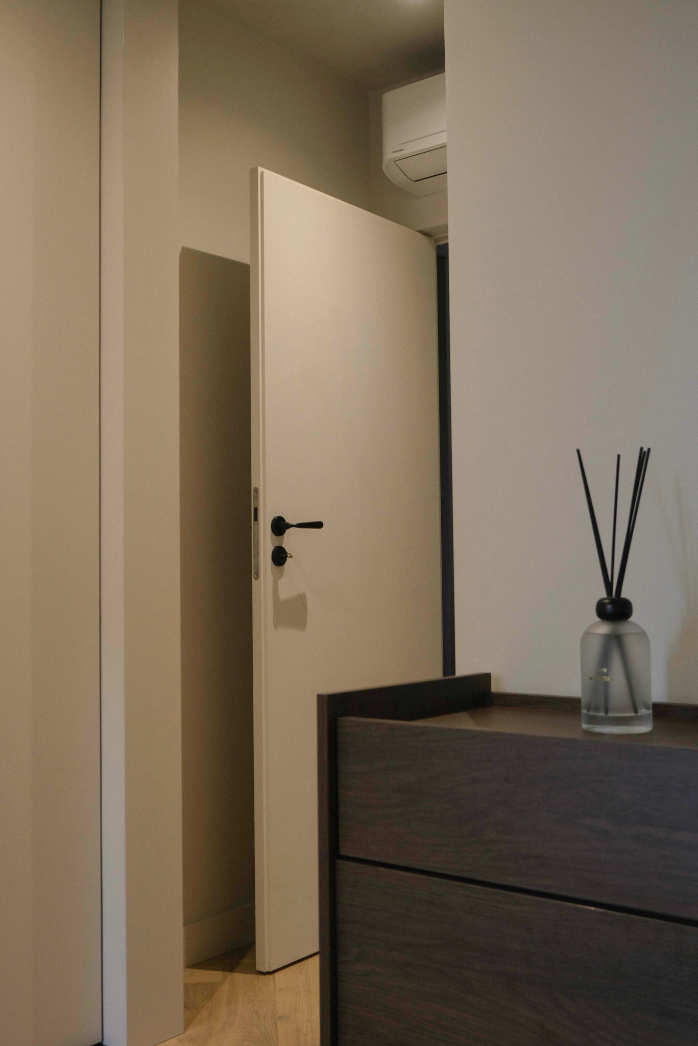 Nowoczesne wnętrze: uchylone białe drzwi z czarną klamką, drewniana komoda z zapachem w szklanym flakonie. Minimalistyczny design, jasne ściany, drewniana podłoga.