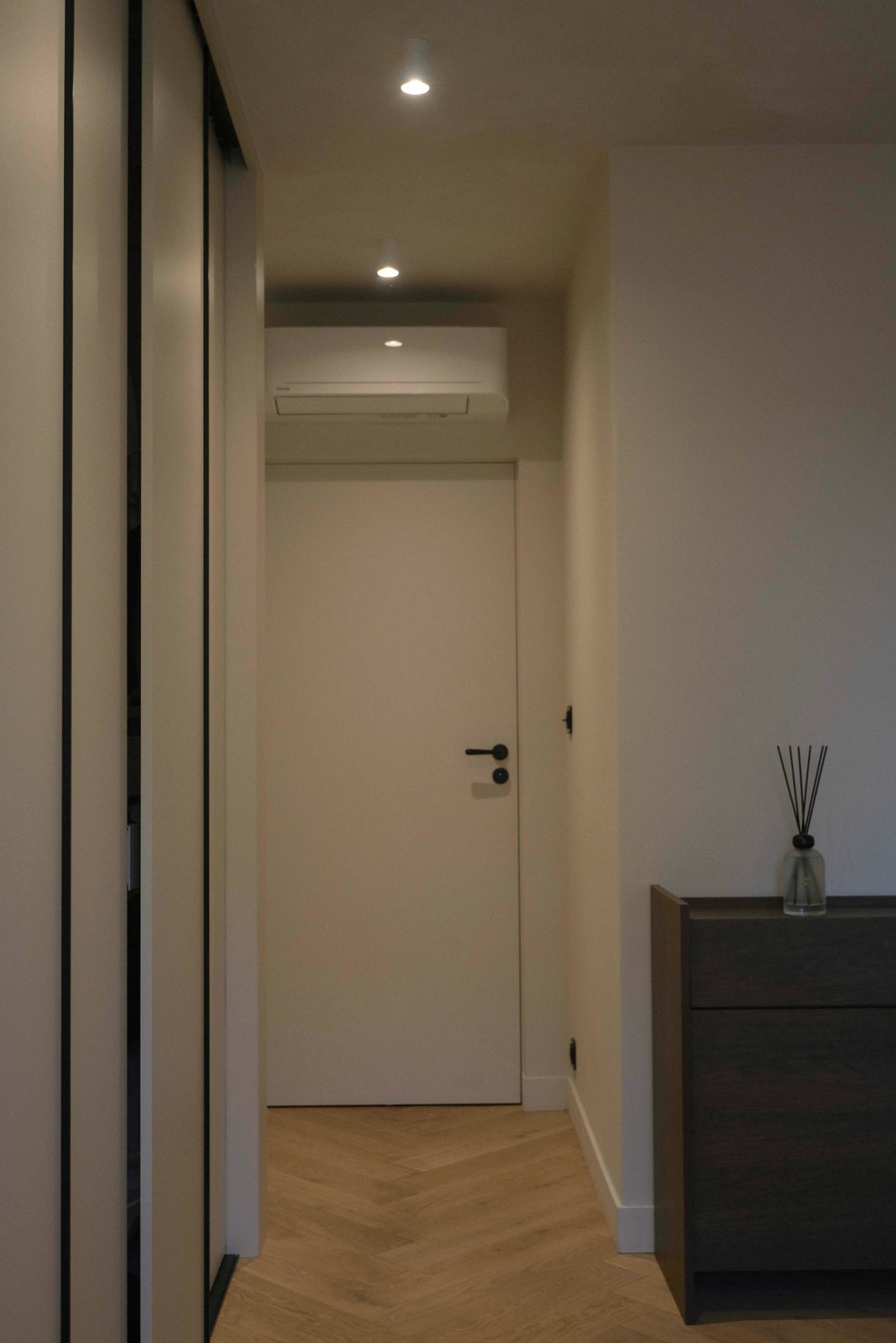 Minimalistyczny korytarz: białe ściany, drzwi i szafa z czarnymi akcentami, parkiet w jodełkę, komoda z dyfuzorem zapachowym. Oświetlenie punktowe.