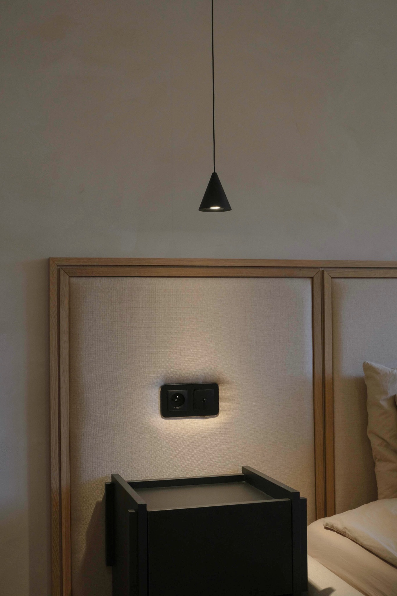 Minimalistyczna sypialnia: lampa stożkowa nad szafką nocną, gniazdko z podświetleniem w wezgłowiu łóżka z beżową tapicerką i drewnianą ramą.