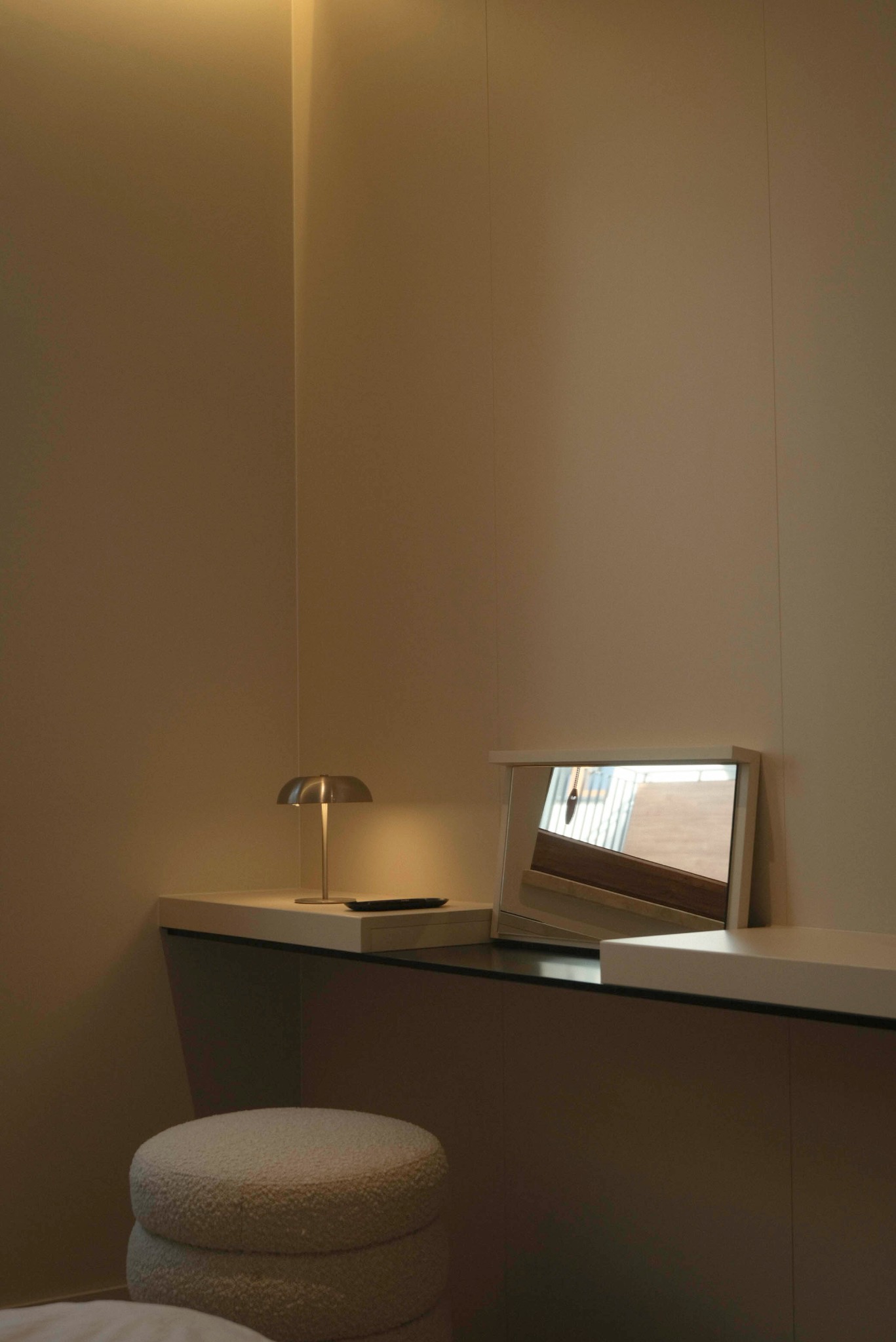 Minimalistyczna aranżacja wnętrza: toaletka z lustrem, lampką i pufą w odcieniach beżu. W lustrze odbija się fragment okna i wnętrza. Ciepłe, stonowane oświetlenie.