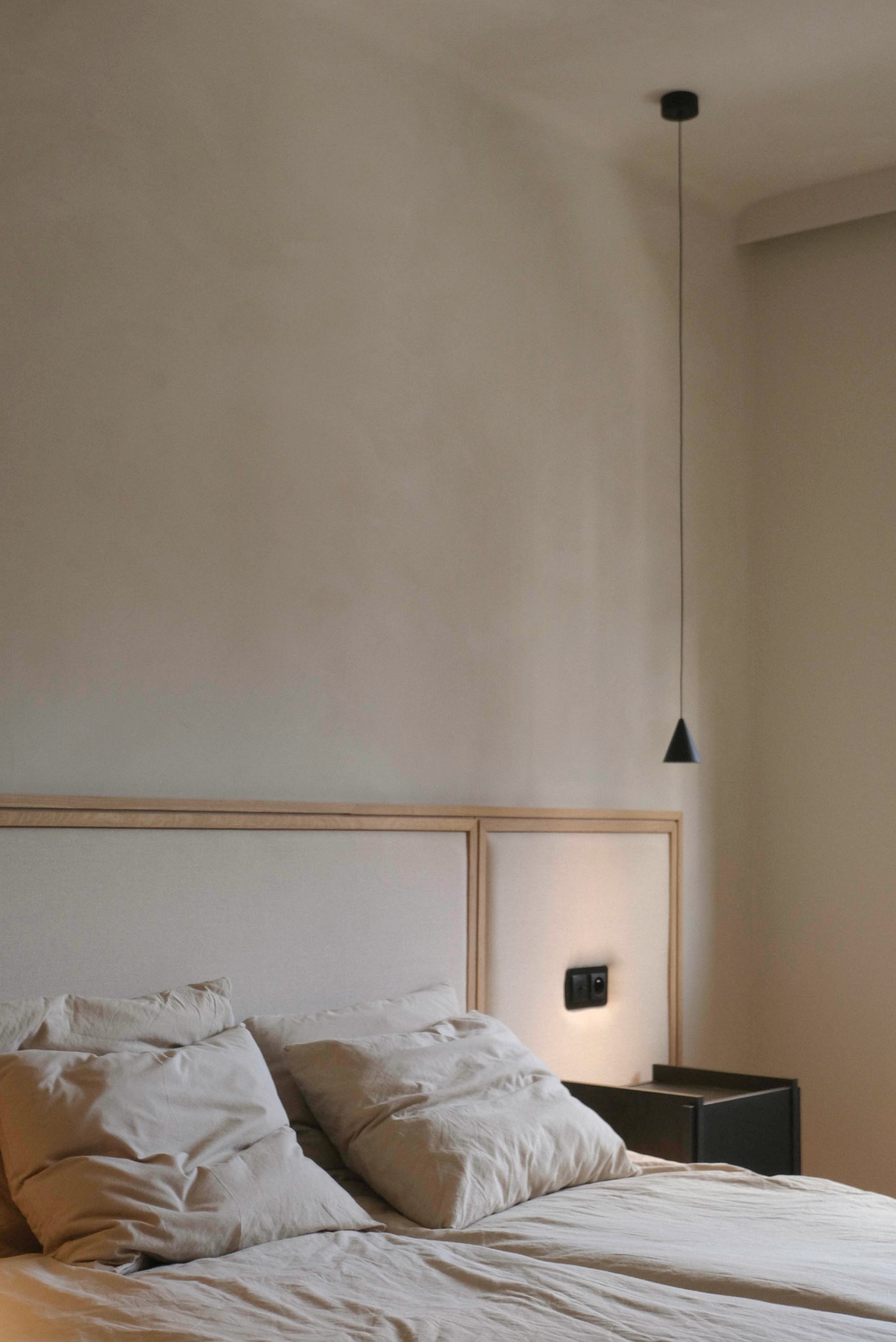 Minimalistyczna sypialnia w beżach: łóżko z tapicerowanym wezgłowiem, lampka wisząca z czarnym kloszem i ciemna szafka nocna. Subtelne oświetlenie tworzy przytulny klimat.