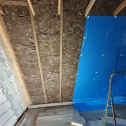 Izolacja poddasza wełną mineralną między krokwiami, częściowo zakryta niebieską folią ochronną, widoczna drabina i fragment podłogi z desek.