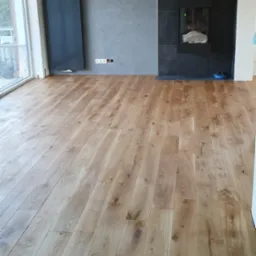 Jasne panele podłogowe ułożone w jodełkę, widoczny grzejnik na ścianie i fragment kominka z płytkami