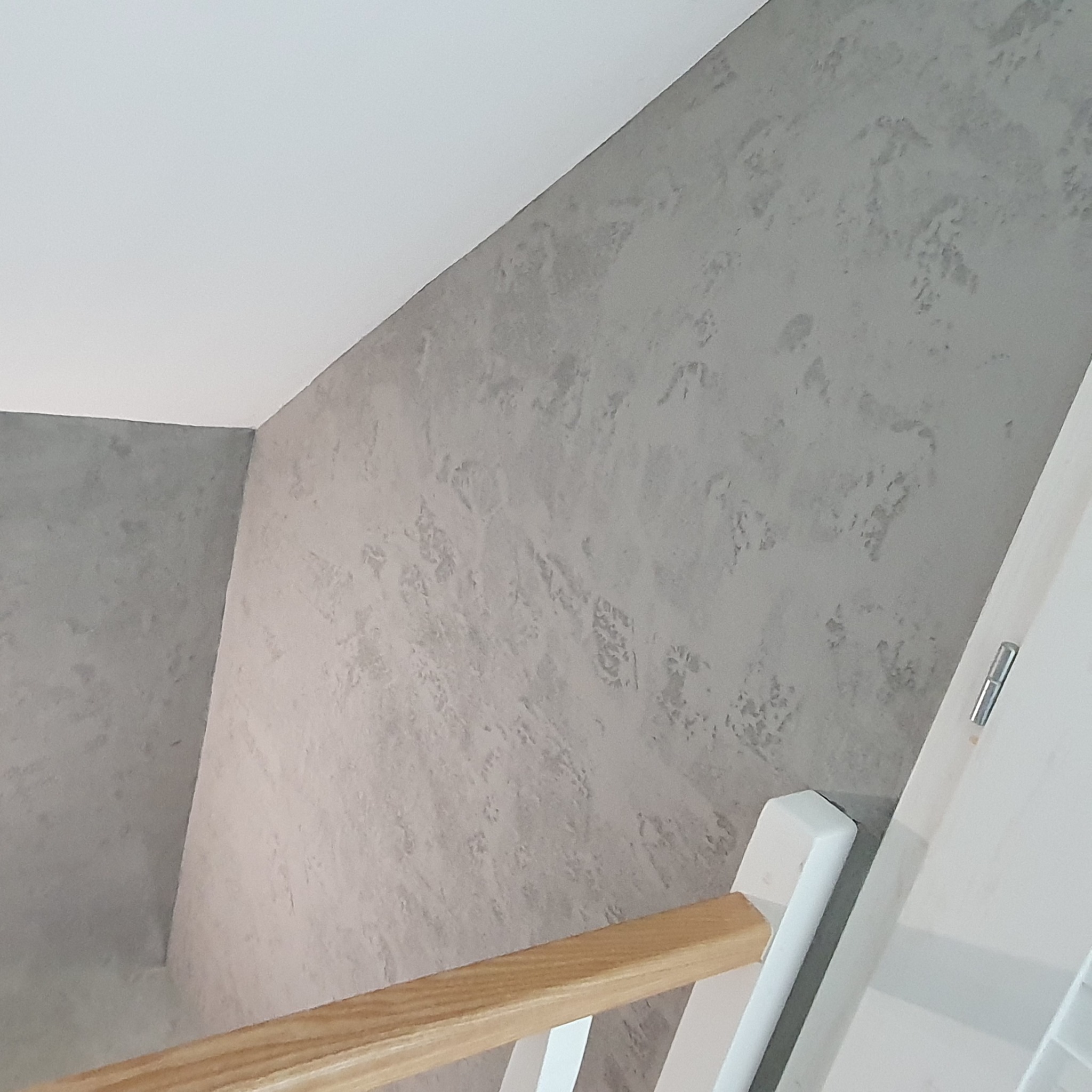 Ściana wykończona szarą, dekoracyjną strukturą przypominającą beton architektoniczny, widoczny fragment białych schodów z drewnianą barierką.