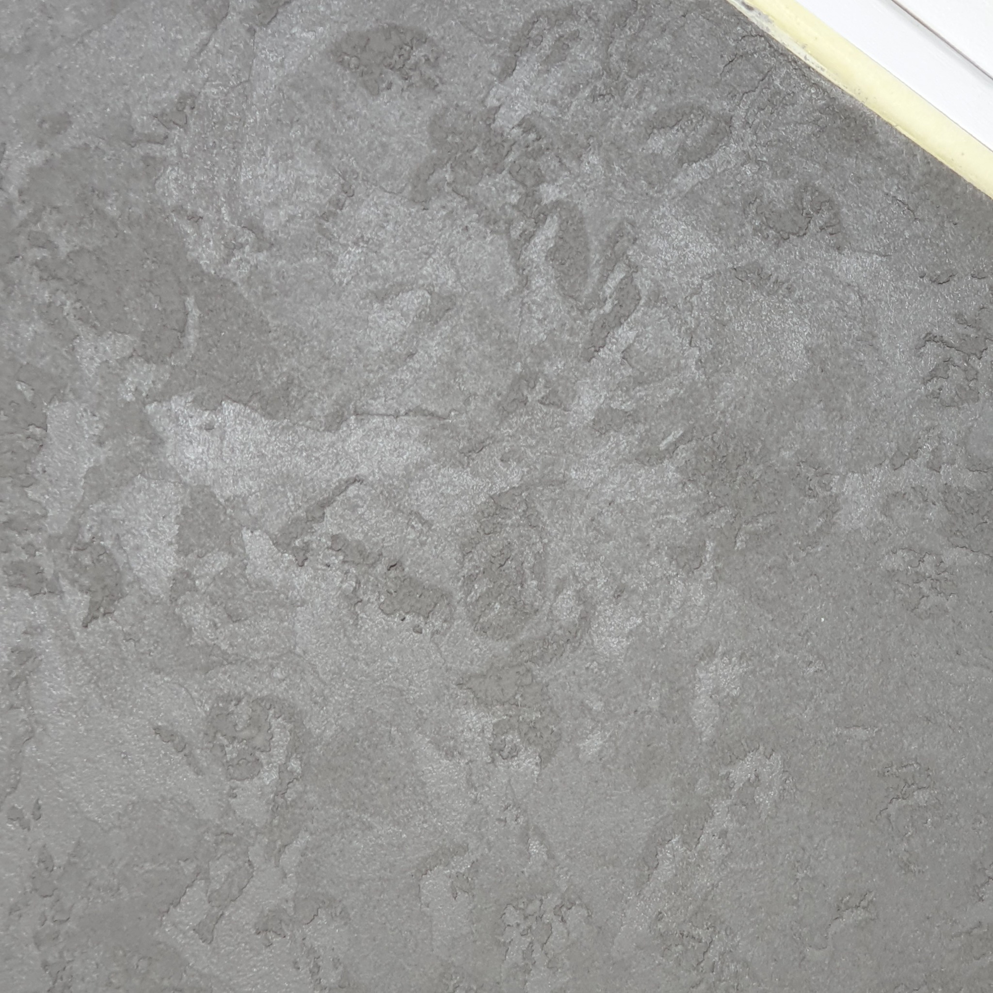 Szara ściana z efektem dekoracyjnym, widoczna struktura i nieregularne plamy koloru, górny róg z widoczną listwą przypodłogową.