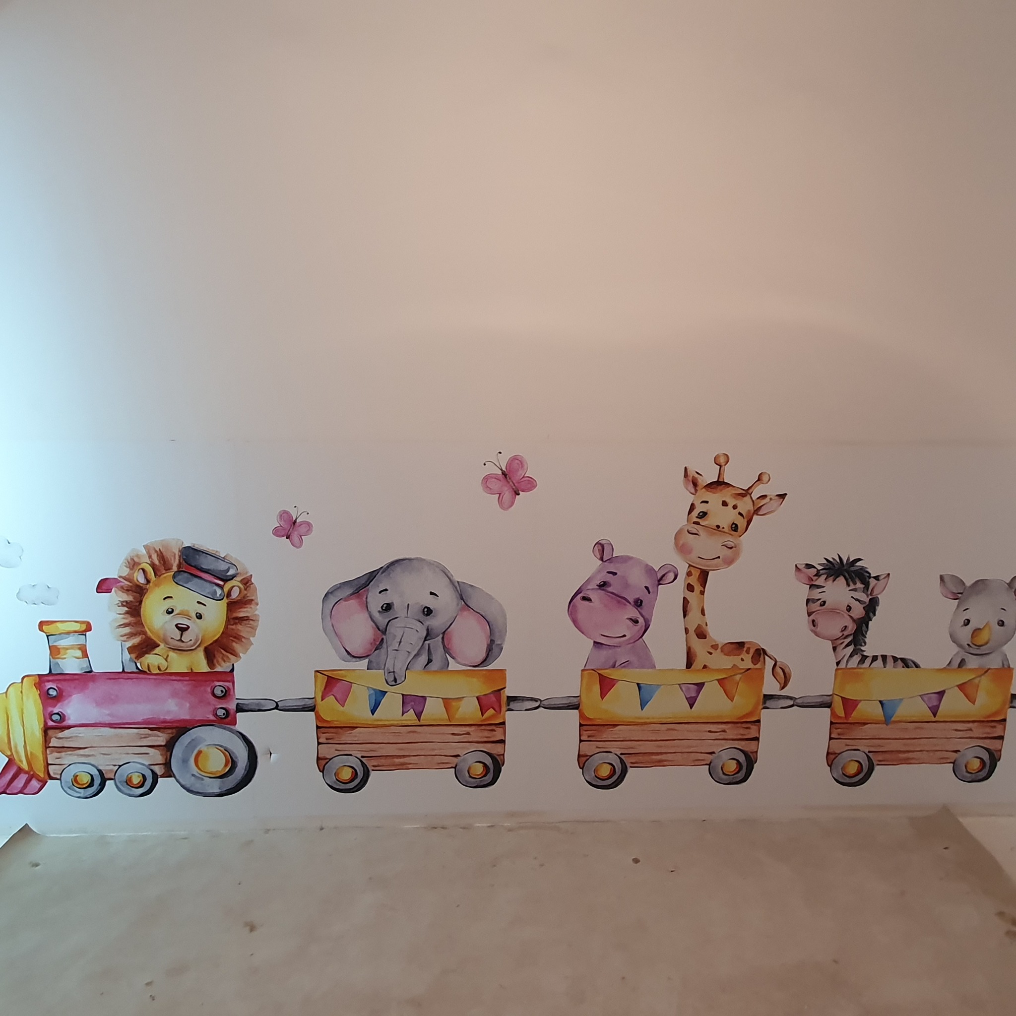Ręcznie malowany na ścianie pokoju dziecięcego pociąg z wagonami, w których siedzą zwierzęta: lew w czapce, słoń, hipopotam, żyrafa, zebra i nosorożec. Nad nimi dwa różowe motyle.