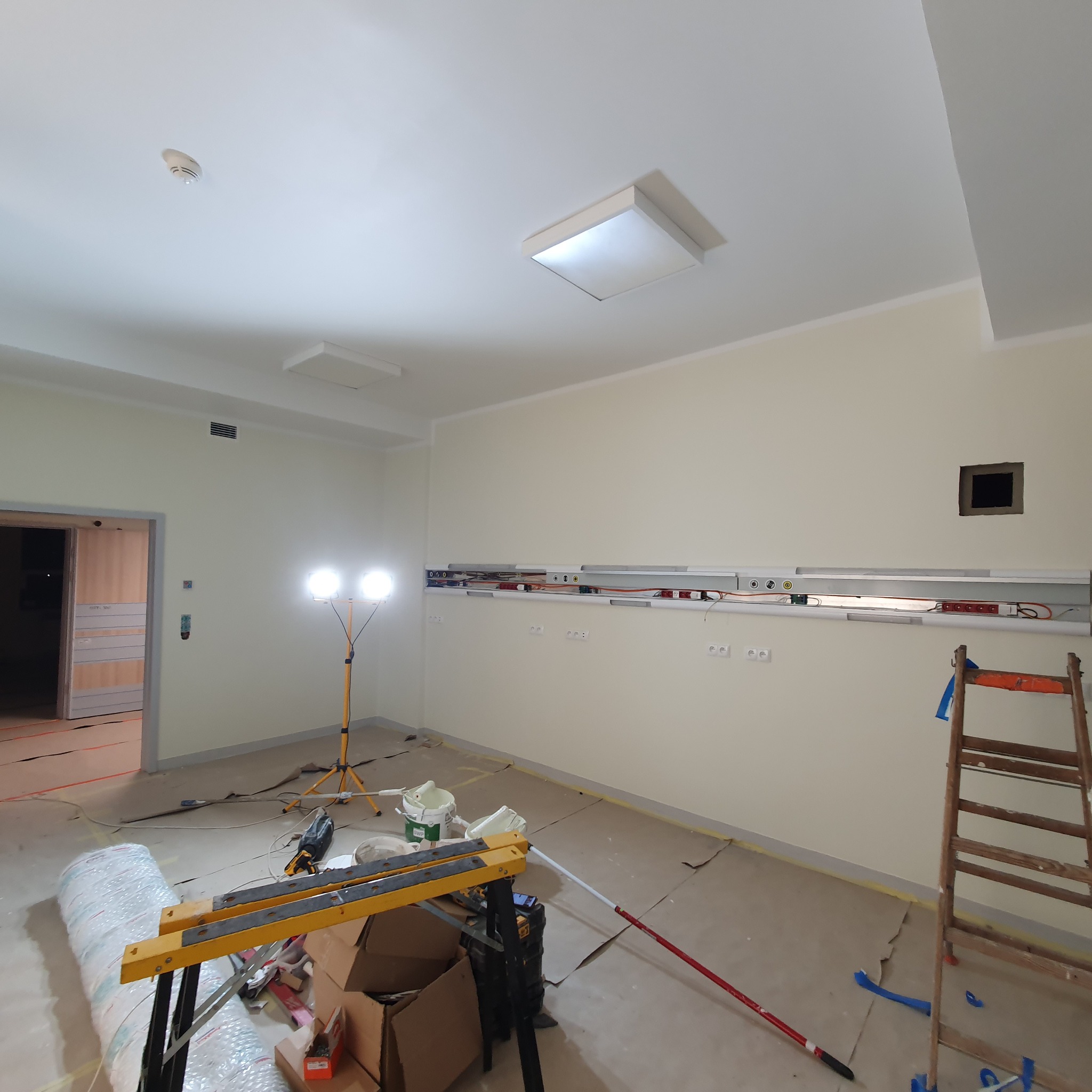 Wnętrze w trakcie remontu: ściany przygotowane do malowania, widoczne poziomice, drabina, lampa halogenowa na statywie, zabezpieczona podłoga kartonem, narzędzia malarskie.