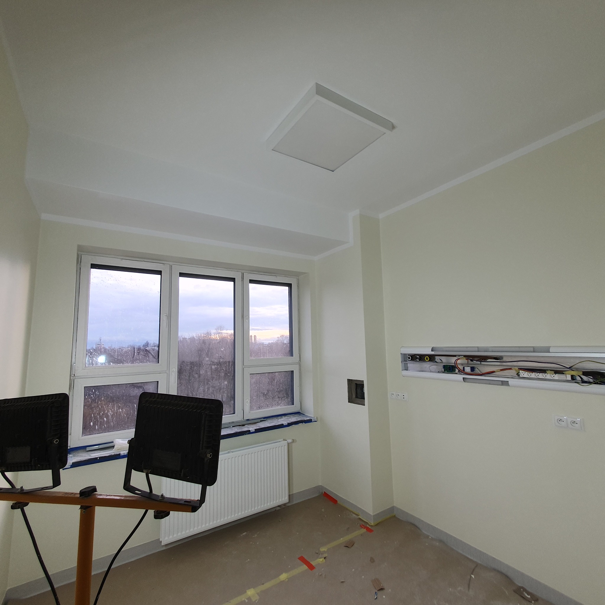 Pokój w trakcie remontu: świeżo pomalowane ściany w odcieniu kremowym, widoczne dwa reflektory na statywie, okno z widokiem na miasto i grzejnik, taśma malarska na podłodze.