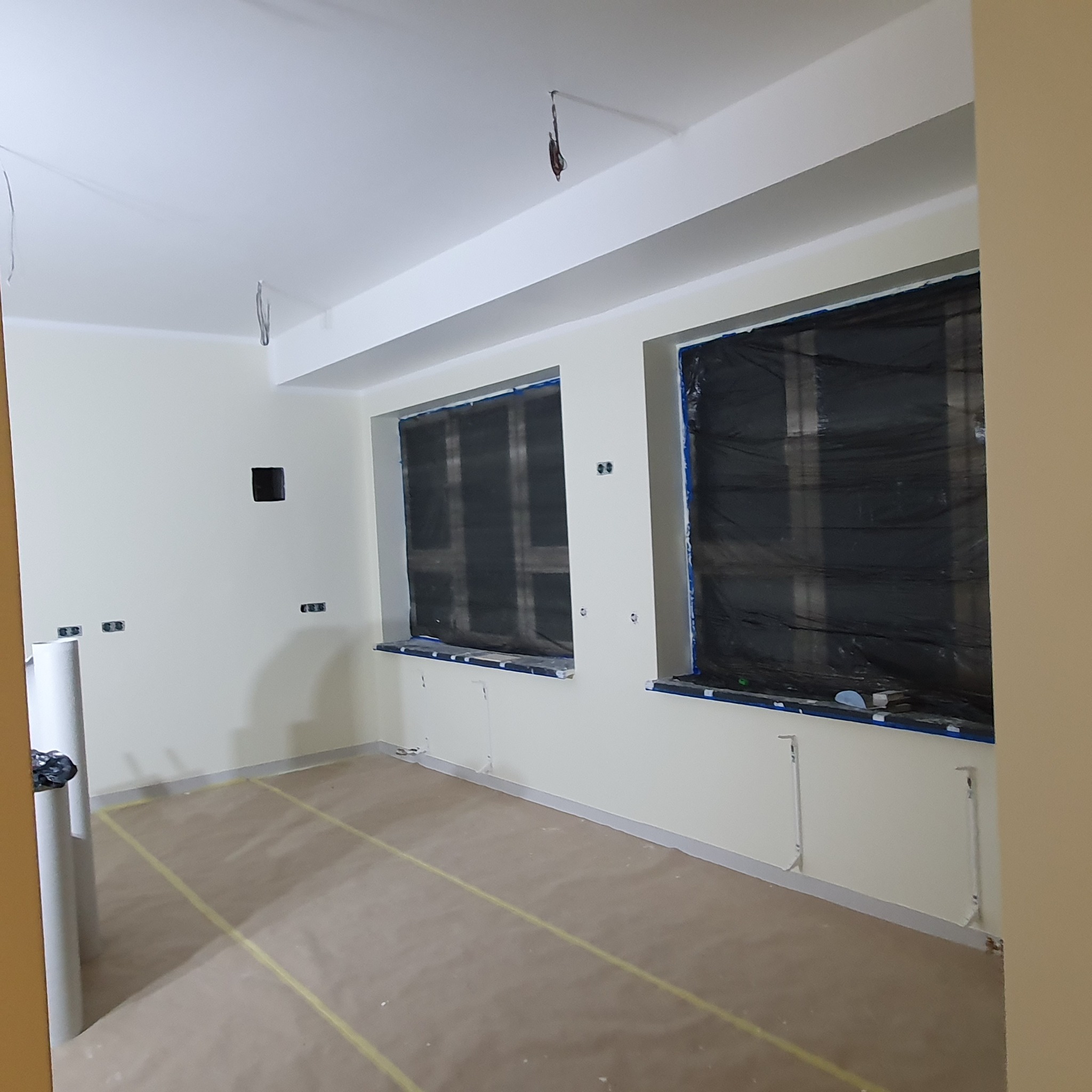 Przygotowane do malowania pomieszczenie z zakrytymi oknami, zabezpieczoną podłogą i świeżo pomalowanymi ścianami na jasny odcień.