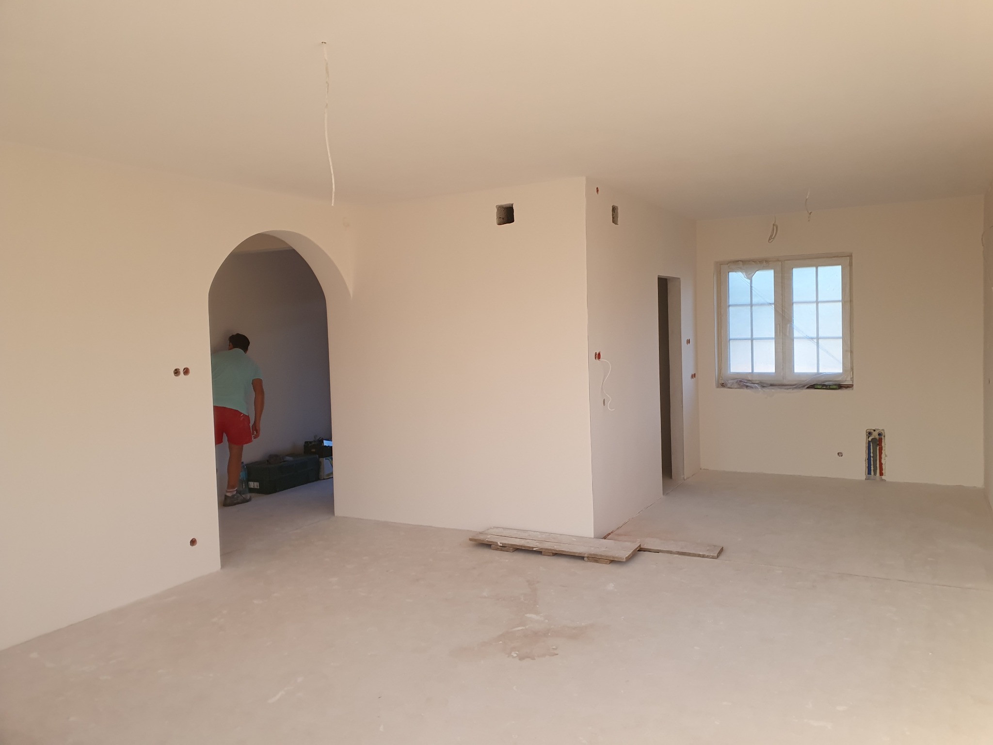 Jasne, puste wnętrze po szpachlowaniu i malowaniu, widoczne ślady po pracach, zabezpieczone okno folią, mężczyzna w oddali w przejściu.