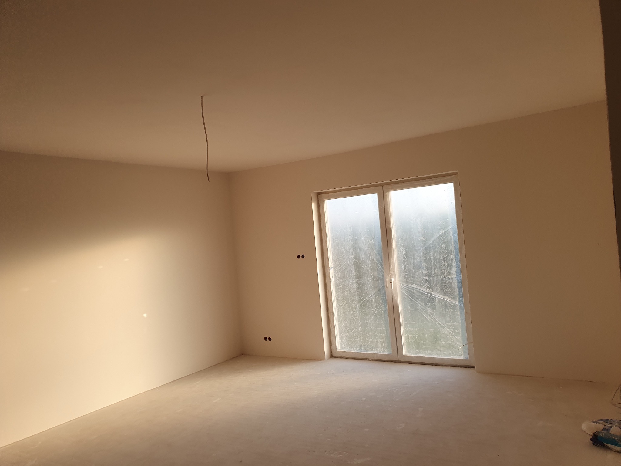 Jasne wnętrze w stanie deweloperskim z gładkimi ścianami i oknem zabezpieczonym folią, widoczny wystający kabel ze ściany.