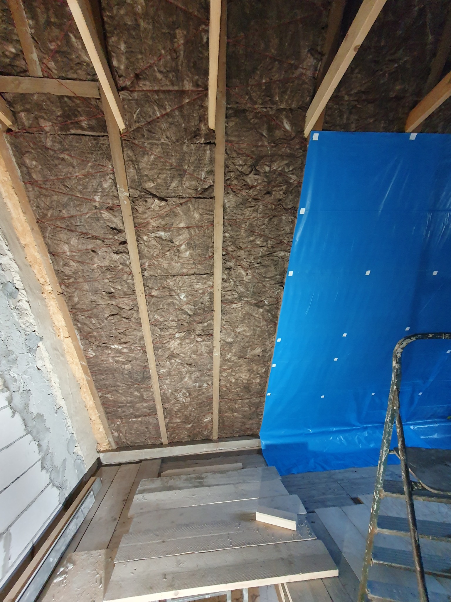 Izolacja poddasza wełną mineralną między krokwiami, częściowo zakryta niebieską folią ochronną, widoczna drabina i fragment podłogi z desek.