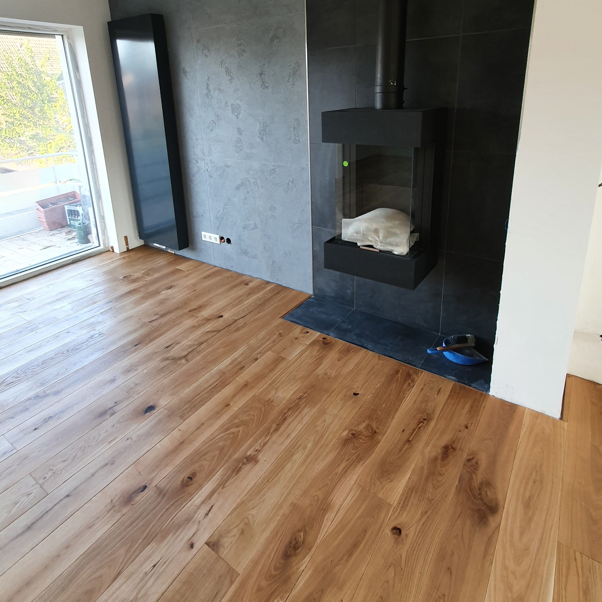 Parkiet z naturalnego drewna ułożony we wnętrzu z kominkiem i grzejnikiem panelowym, widoczne gniazdka elektryczne w ścianie.