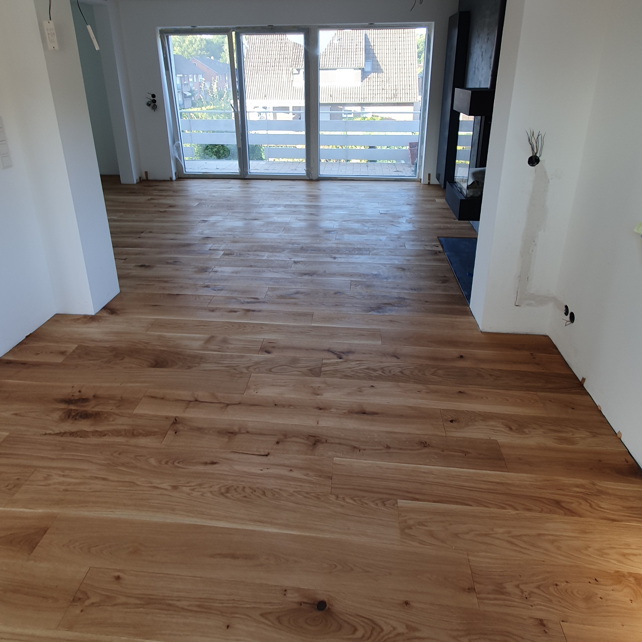 Nowo ułożona podłoga z paneli drewnianych w jasnym odcieniu dębu, widok na duże okno balkonowe i fragment ściany z kominkiem, widoczne niedokończone prace wykończeniowe.