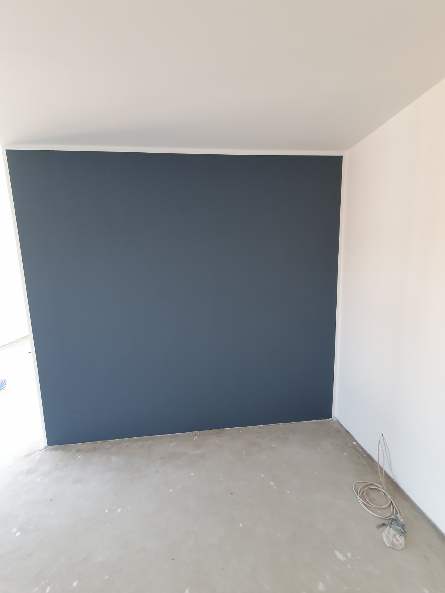 Wnętrze pokoju po malowaniu jednej ściany na ciemny grafit, widoczne kable elektryczne przy podłodze, surowa podłoga przed wykończeniem.