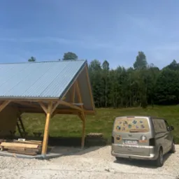 Drewniana wiata z metalowym dachem w trakcie budowy, obok zaparkowany samochód dostawczy z reklamą agroturystyki w tle zielone drzewa i błękitne niebo.