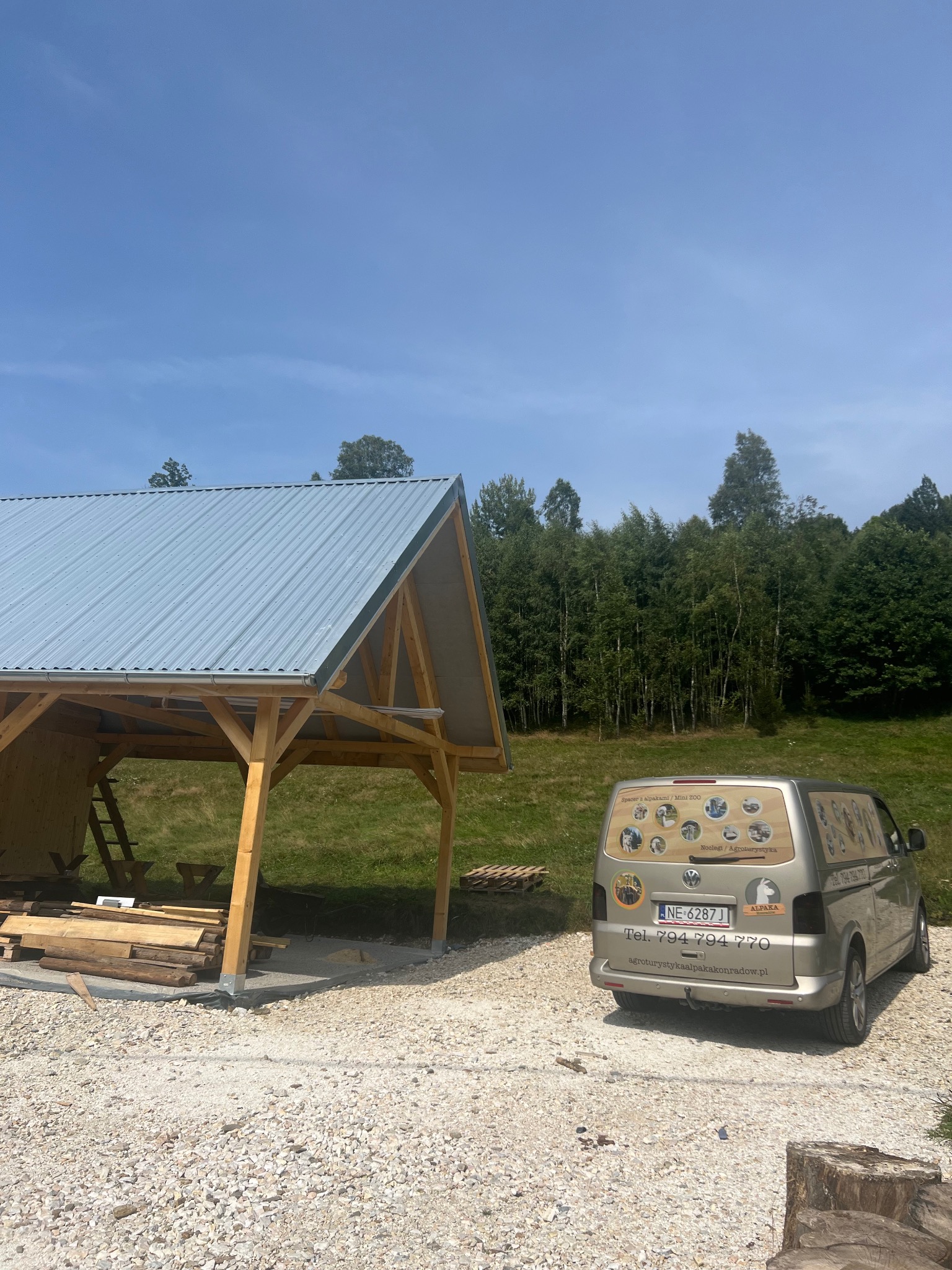 Drewniana wiata z metalowym dachem w trakcie budowy, obok zaparkowany samochód dostawczy z reklamą agroturystyki w tle zielone drzewa i błękitne niebo.