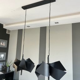 ElektroCare - Dwa czarne, nowoczesne klosze lamp wiszących z geometrycznymi elementami, zamontowane na suficie w pokoju z szaro-białymi pasami na ścianie.
