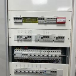 Rozdzielnica elektryczna z zabezpieczeniami nadprądowymi, wyłącznikami r&oacute;żnicowoprądowymi i ogranicznikiem przepięć w białej obudowie, widoczne okablowanie, etykiety opisujące obwody.