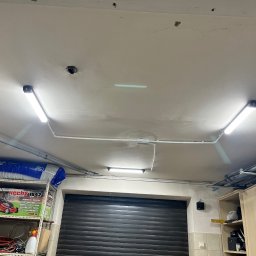 ElektroCare - Sufit garażu z zamontowanymi, liniowymi lampami LED, widoczne rury instalacyjne i półki z różnymi przedmiotami, w tym opakowaniem kosiarki Hecht 1842.