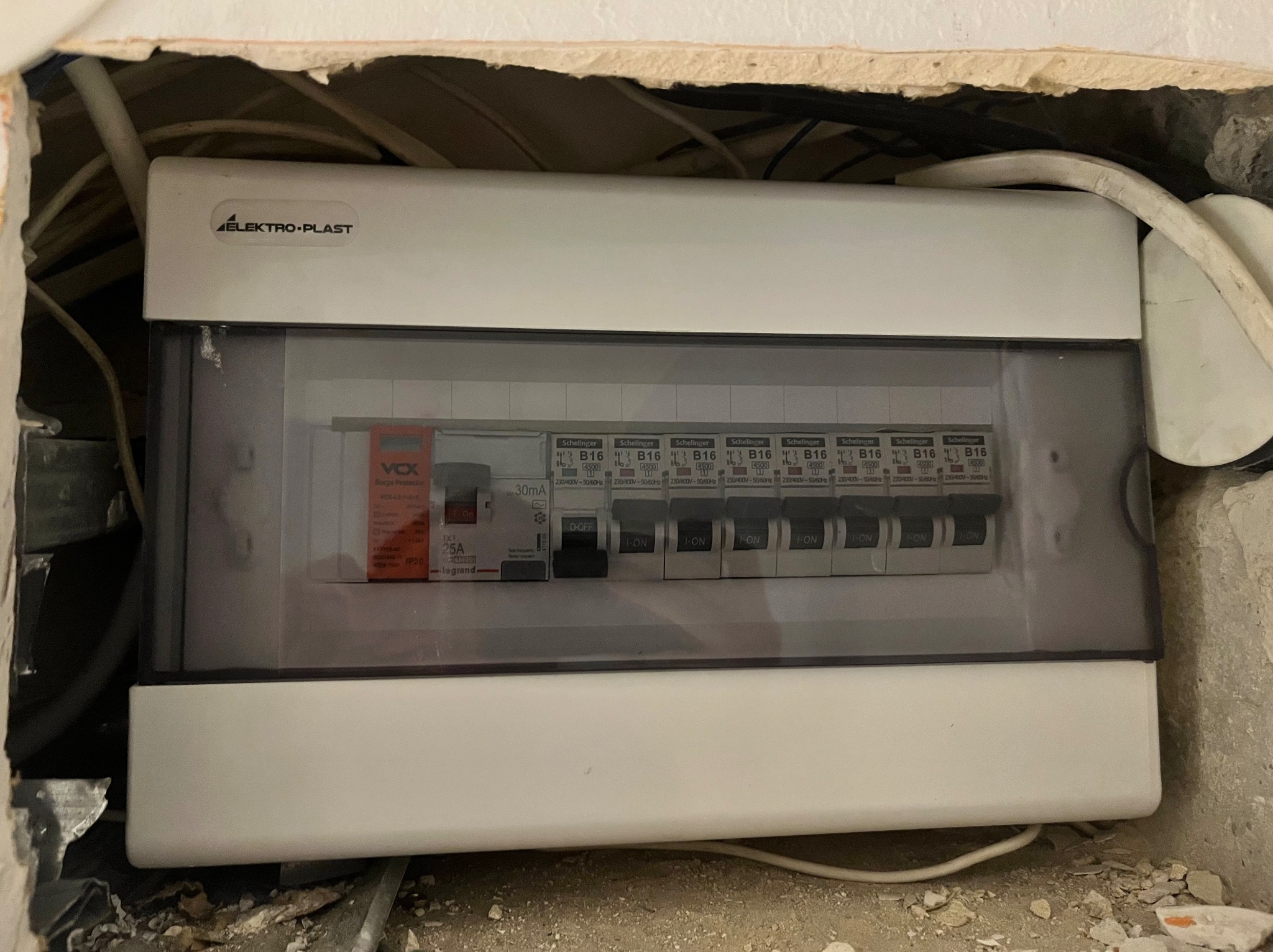 Rozdzielnica elektryczna Elektro-Plast z zainstalowanymi bezpiecznikami Schelinger B16 i ogranicznikiem przepięć VOX, zamontowana w surowym otoczeniu, widoczne kable i fragmenty gruzu.