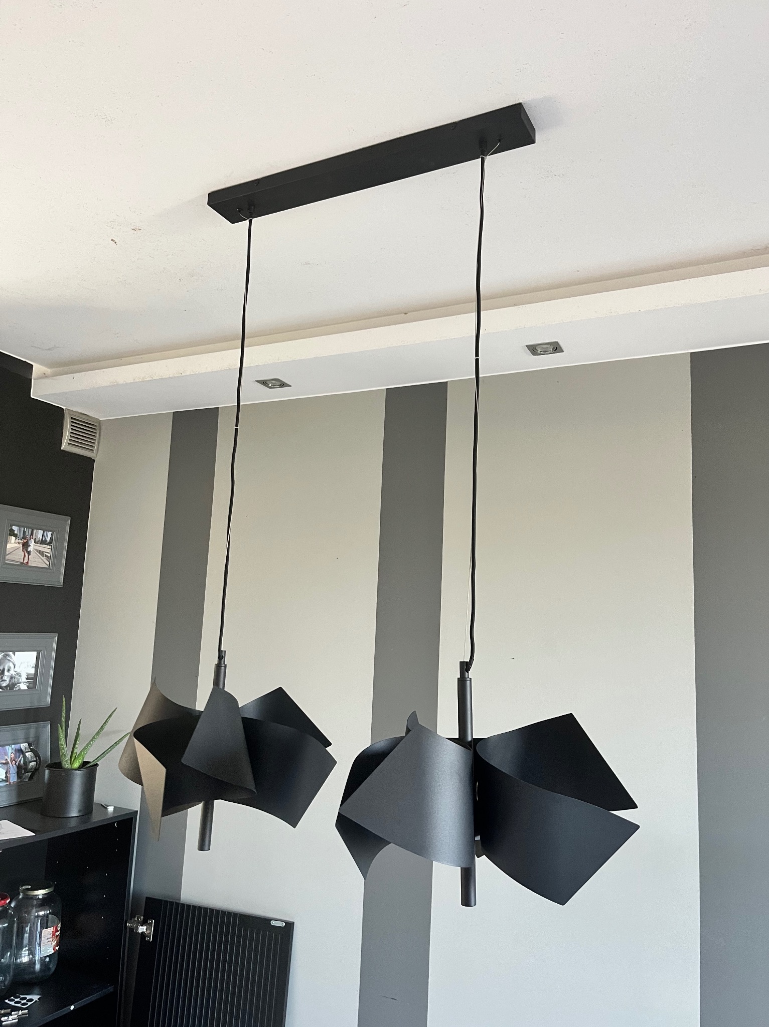 Dwa czarne, nowoczesne klosze lamp wiszących z geometrycznymi elementami, zamontowane na suficie w pokoju z szaro-białymi pasami na ścianie.