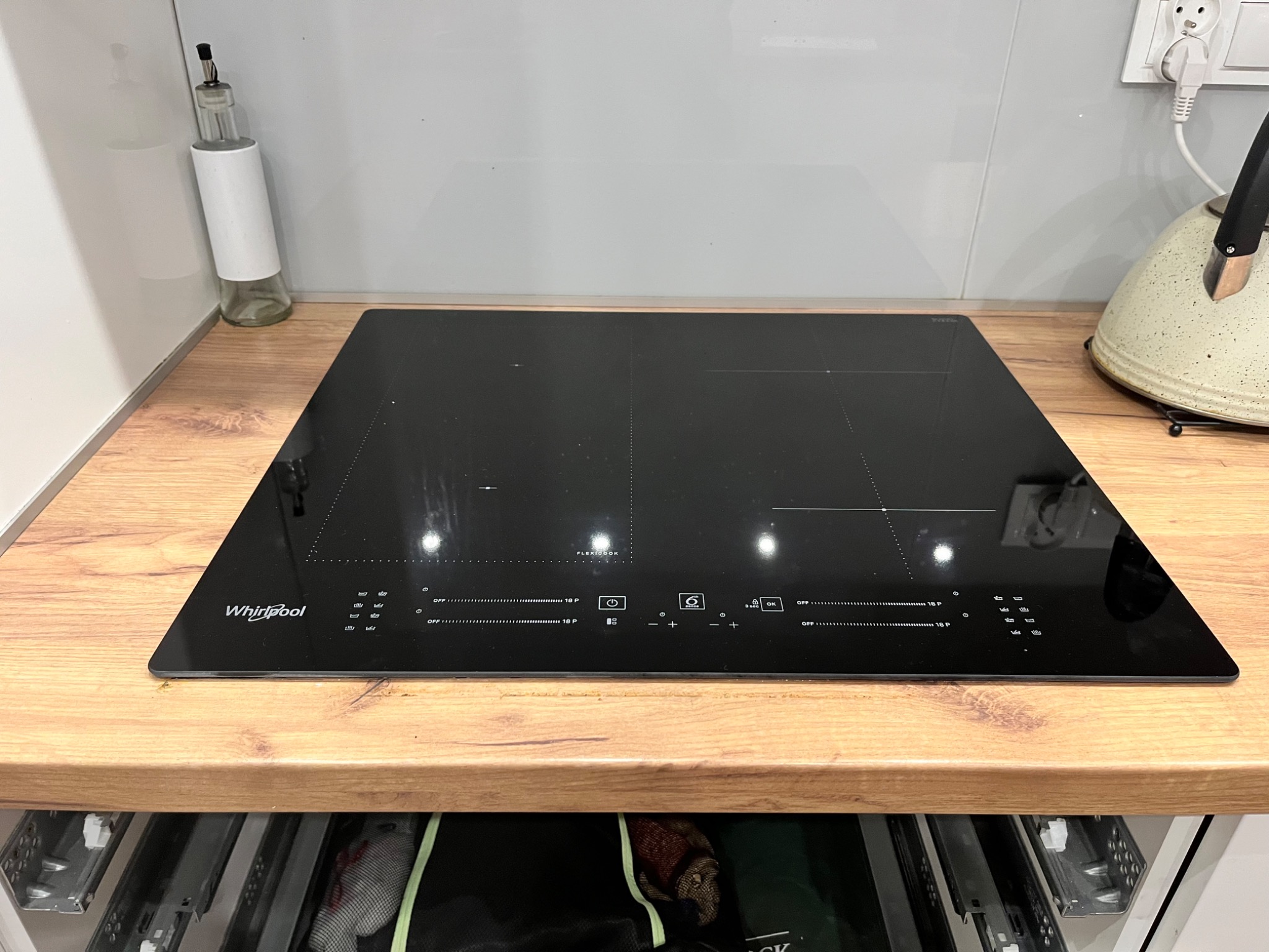 Czarna płyta indukcyjna marki Whirlpool na drewnianym blacie kuchennym, z widocznymi elementami sterowania dotykowego i naczyniem na olej obok.