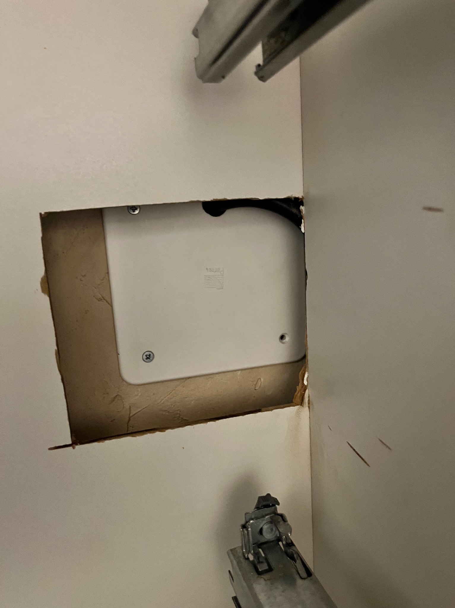 Otwór w ścianie z widoczną białą puszką instalacyjną zabezpieczoną dwoma śrubami, obok widoczny fragment mocowania szuflady