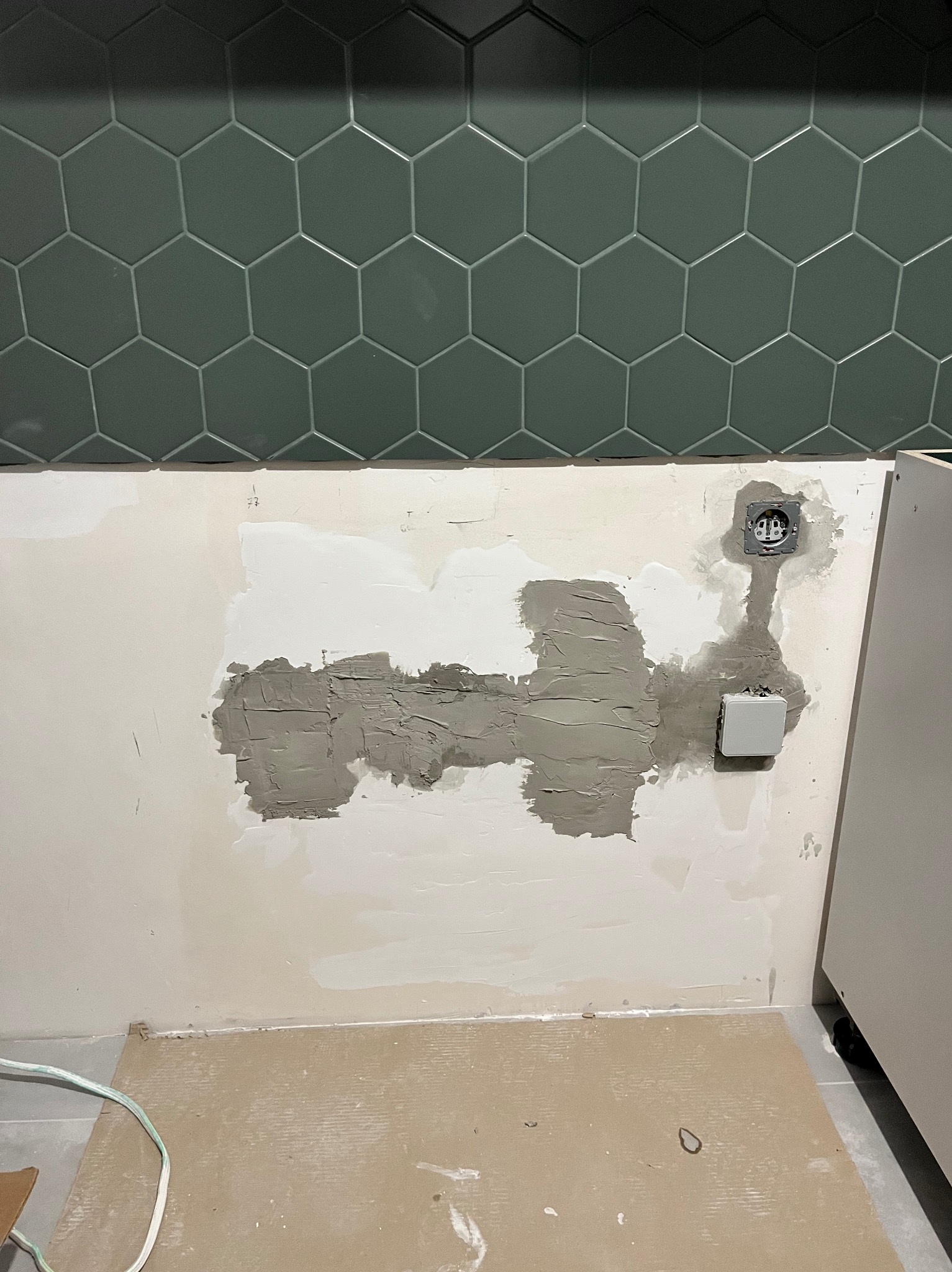 Przygotowanie ściany pod płytki heksagonalne: szpachlowanie nierówności wokół gniazdka elektrycznego i włącznika, widoczne kable i fragment szafki.