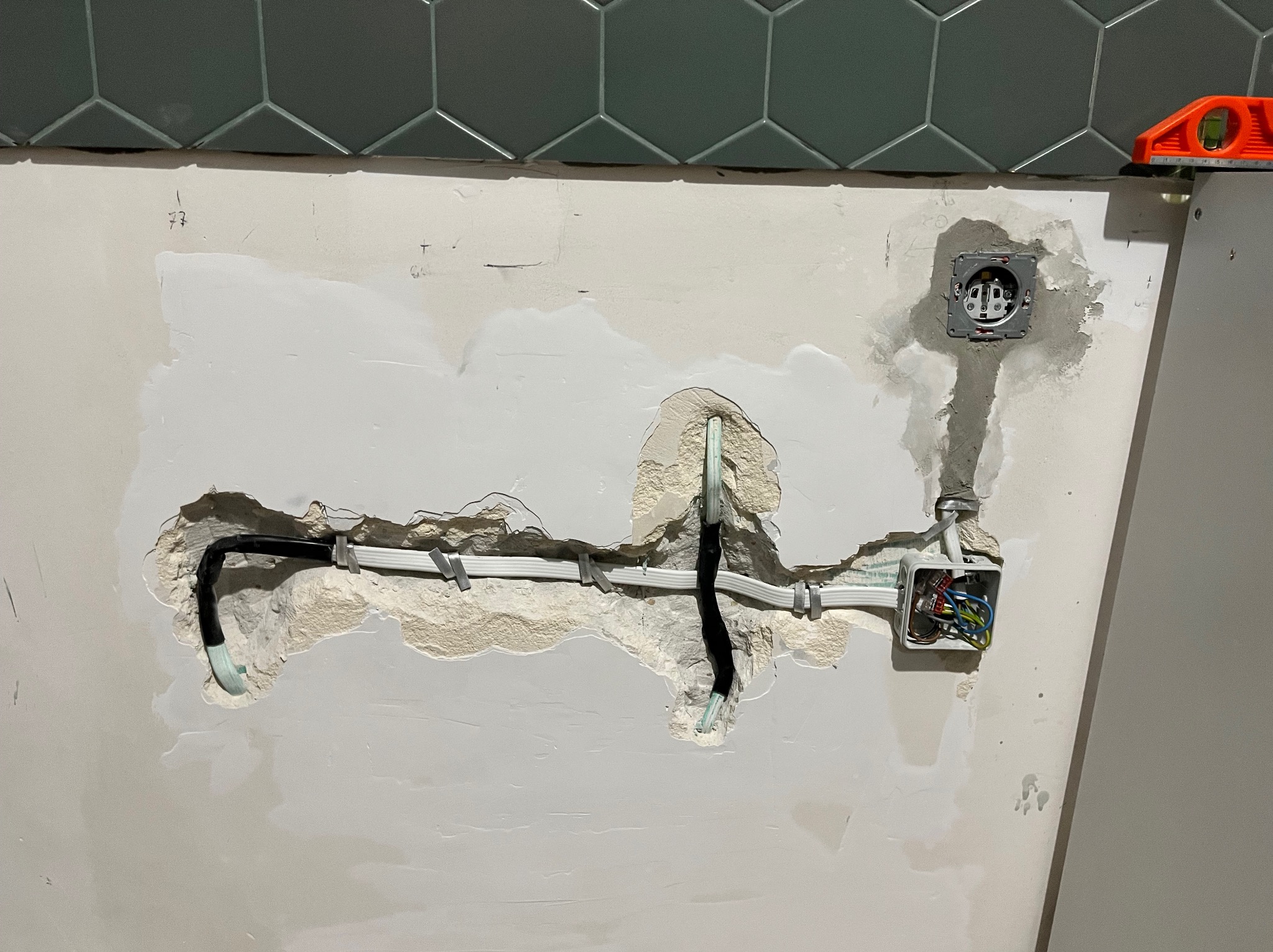 Fragment ściany z widocznymi przewodami elektrycznymi w trakcie prac instalacyjnych, puszka pod gniazdko i otwarta puszka połączeniowa z kolorowymi kablami, poziomica na krawędzi ściany, tło...