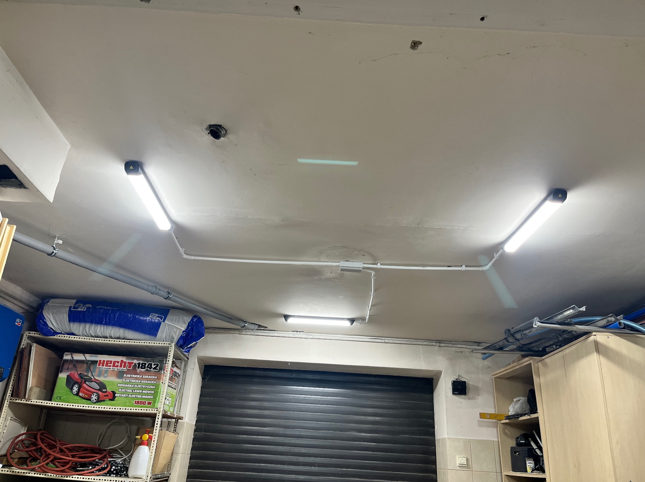 Sufit garażu z zamontowanymi, liniowymi lampami LED, widoczne rury instalacyjne i półki z różnymi przedmiotami, w tym opakowaniem kosiarki Hecht 1842.