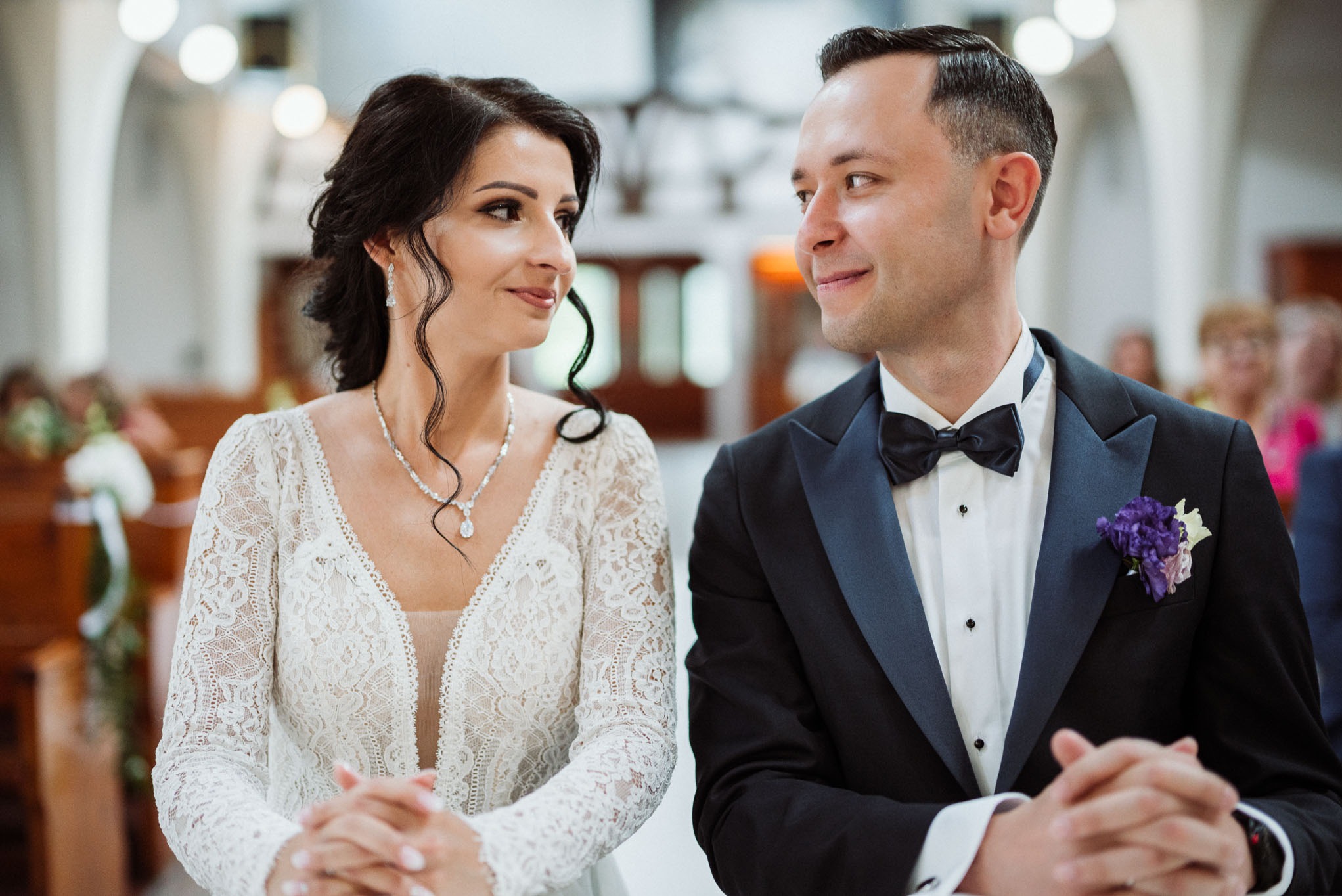 Uśmiechnięta para młoda w kościele: panna młoda w koronkowej sukni z długim rękawem i dekoltem w serek, pan młody w czarnym smokingu z fioletowym butonierem, oboje z dłońmi złożonymi przed sobą.
