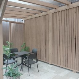 Usługi stolarskie - Zadaszona przestrzeń z drewnianą konstrukcją, pionowymi lamelami tworzącymi ścianę, oraz zestawem mebli ogrodowych: kwadratowy stół i cztery krzesła. Rośliny w donicach dodają naturalnego akcentu.