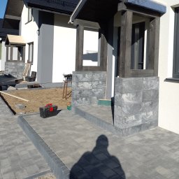 Usługi stolarskie - Budowa wejścia do domu z szarej kostki brukowej i drewnianej konstrukcji zadaszenia, widoczna skrzynka narzędziowa i miotła, w tle przygotowany teren.