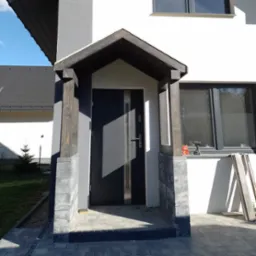 Zadaszenie nad wejściem do domu z drewnianymi słupami i szarą stolarką okienną; widoczny wiertarko-wkrętak na parapecie okiennym.