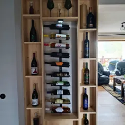 Drewniana szafka na wino z półkami i metalowym uchwytem na butelki, prezentująca różnorodne butelki wina i kieliszki, z widokiem na salon w tle.