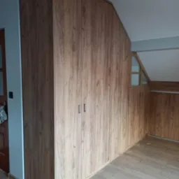 Wysoka zabudowa szafy z drewnopodobną okleiną, dopasowana do skosu poddasza, z widocznymi metalowymi uchwytami i fragmentem podłogi z paneli.