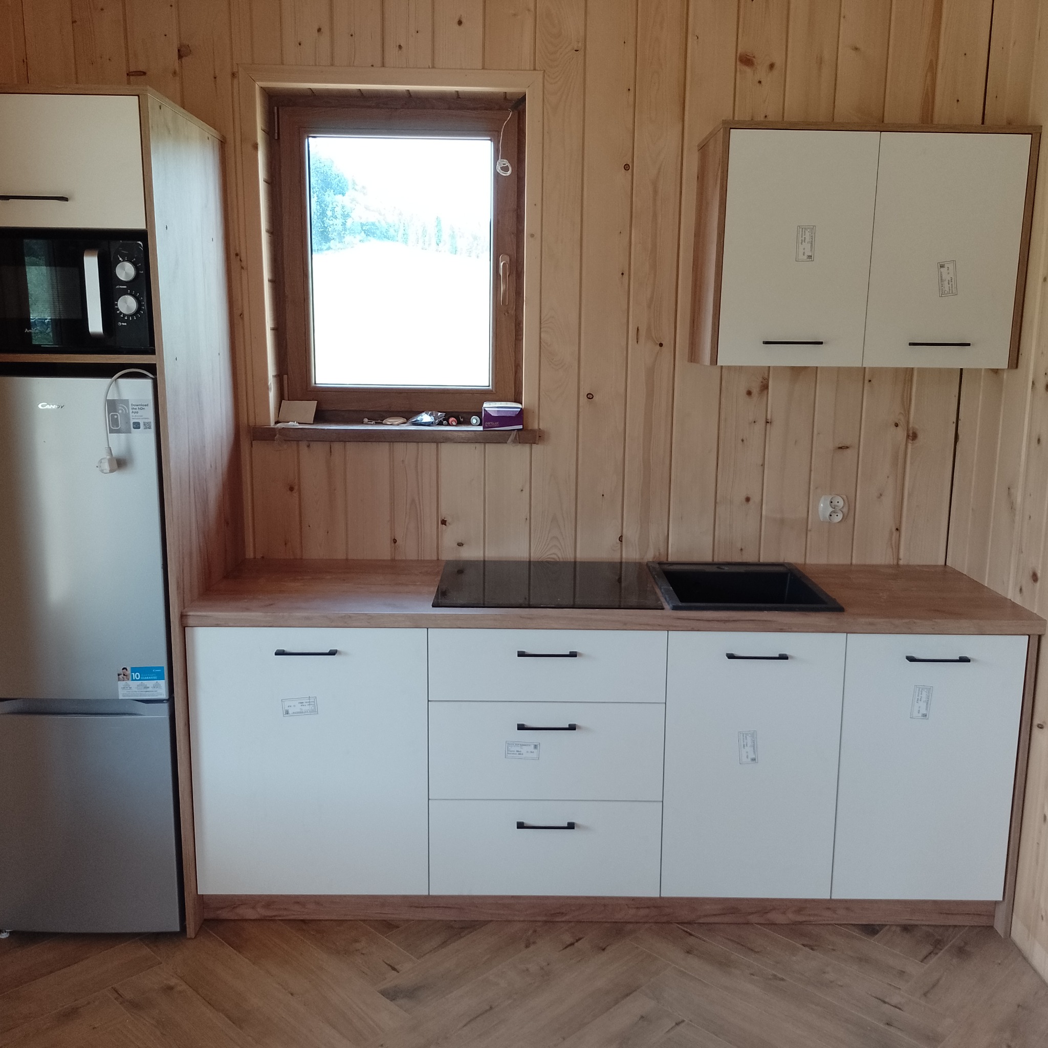 Wnętrze drewnianego domku z zabudową kuchenną: białe szafki z czarnymi uchwytami, drewniany blat z czarną płytą indukcyjną i zlewozmywakiem, lodówka w zabudowie, okno z drewnianą ramą i drewniane...