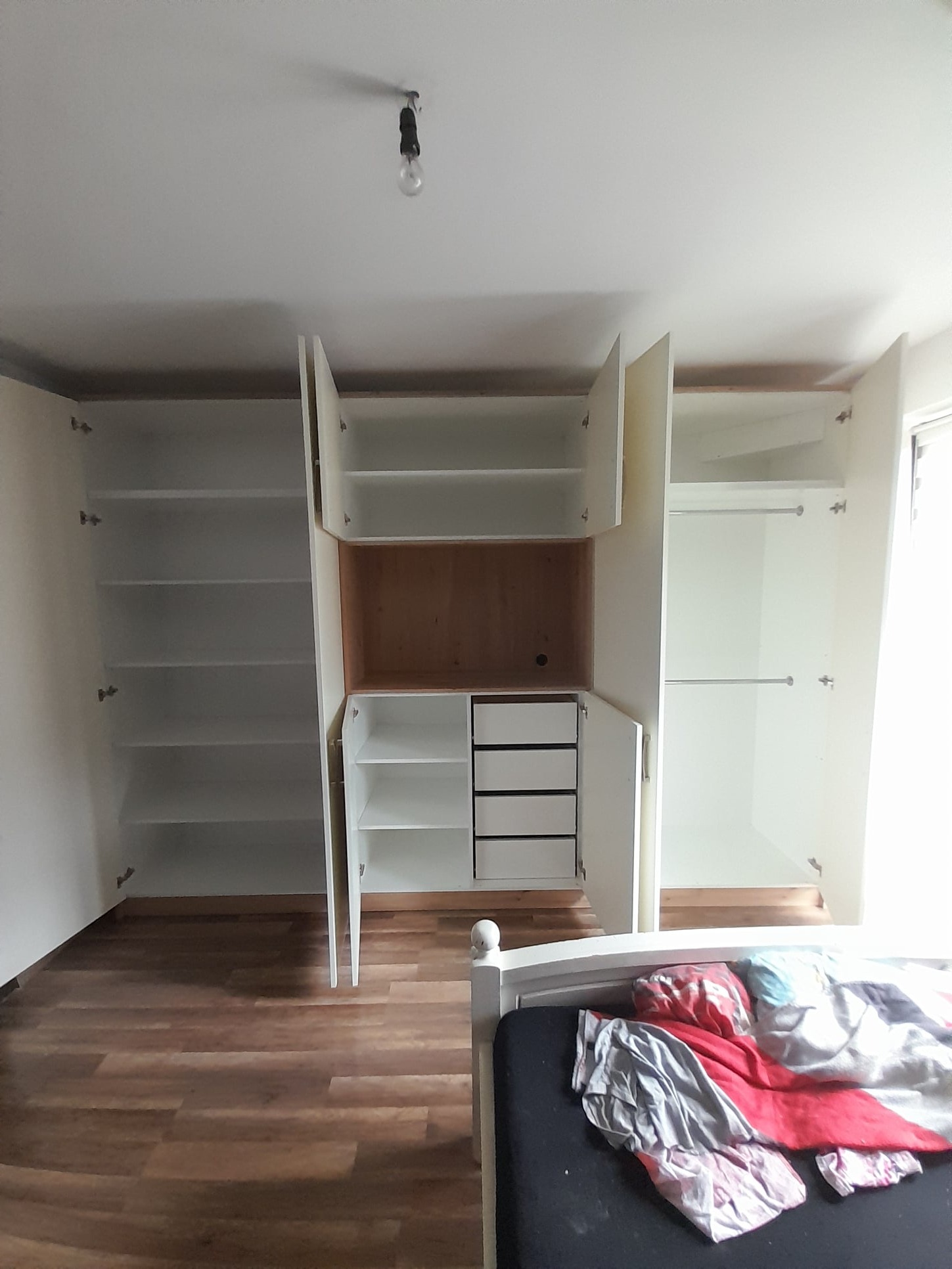 Wnętrze sypialni z dużą, białą szafą wnękową z otwartymi drzwiami, ukazującą półki i szuflady, widok na drewnianą podłogę i fragment łóżka z pościelą.