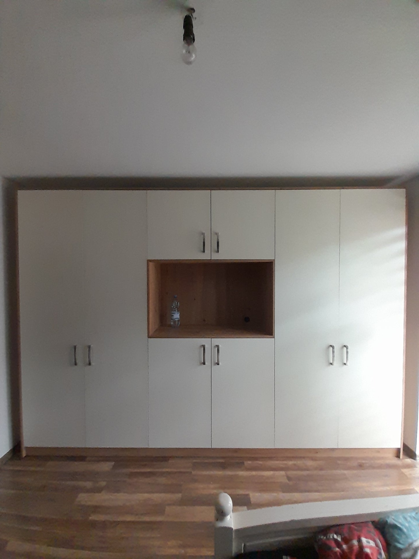 Biała, zabudowana szafa z drewnianym wnęką na środku, na której stoi butelka wody, widoczna na tle parkietu i żarówki na suficie.