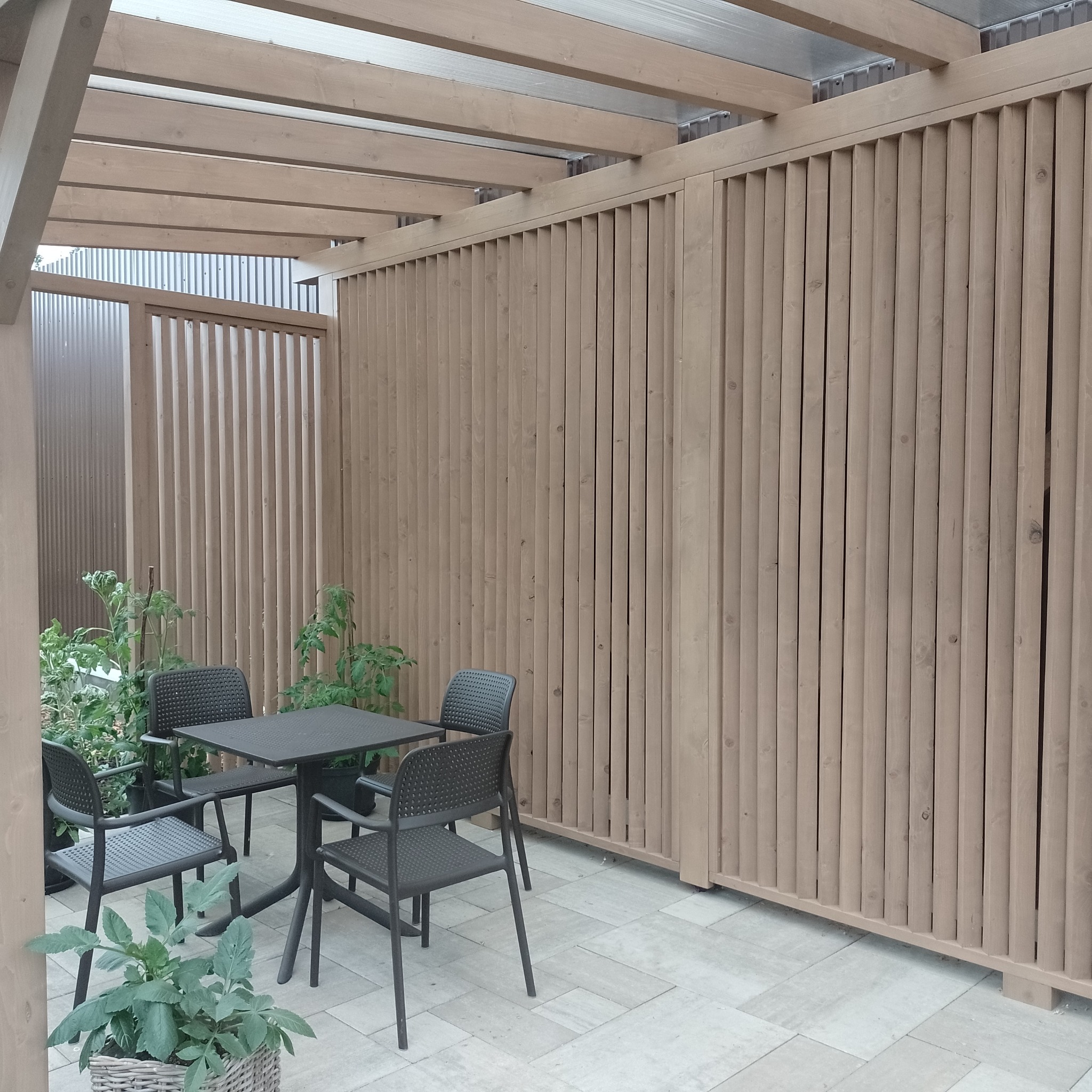 Zadaszona przestrzeń z drewnianą konstrukcją, pionowymi lamelami tworzącymi ścianę, oraz zestawem mebli ogrodowych: kwadratowy stół i cztery krzesła. Rośliny w donicach dodają naturalnego akcentu.