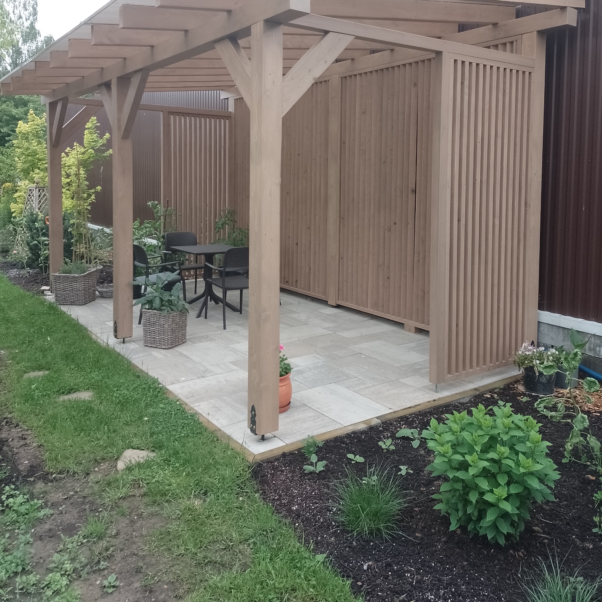 Drewniana pergola z ażurowymi ścianami bocznymi, wyposażona w czarny stół i krzesła, otoczona zielenią i brukowanym tarasem.