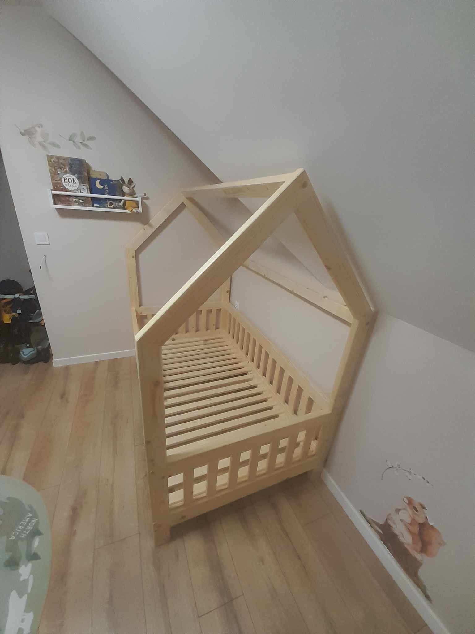 Drewniane łóżko dziecięce w kształcie domku, z drewnianym stelażem, ustawione w pokoju na poddaszu z drewnianą podłogą, obok naklejki ze zwierzętami na ścianie i półki z książkami.
