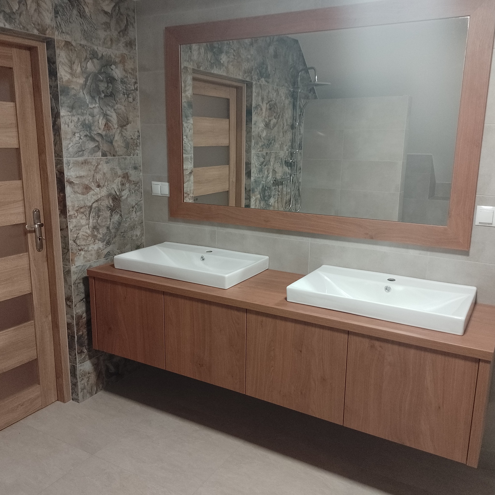 Nowoczesna łazienka z drewnianą szafką pod umywalki nablatowe i dużym lustrem w drewnianej ramie, widoczna również drewniana stolarka drzwiowa i dekoracyjne płytki ścienne z motywem roślinnym.