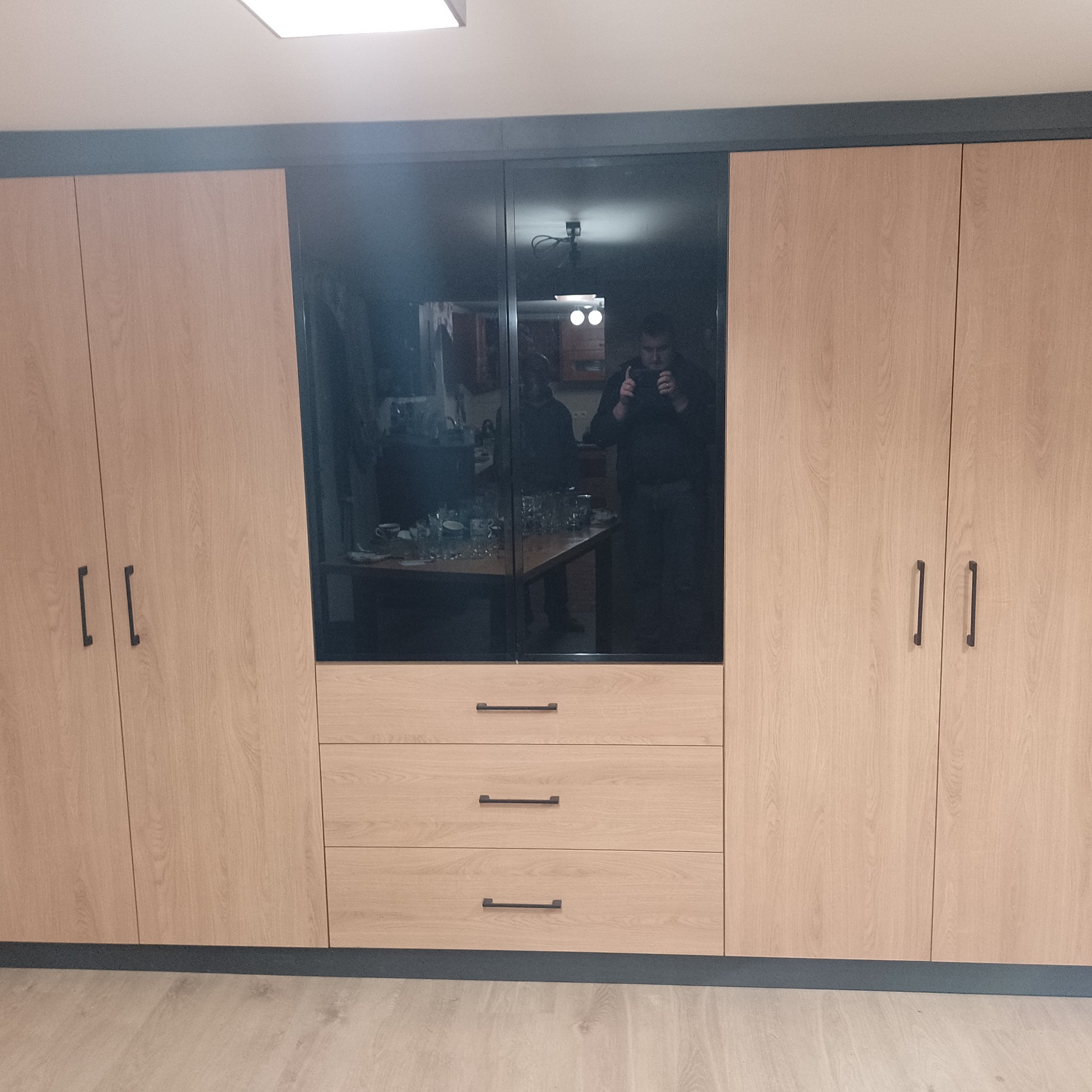 Duża szafa z jasnego drewna z czarnymi, lustrzanymi drzwiami pośrodku, w których odbija się osoba robiąca zdjęcie telefonem oraz stół z naczyniami.