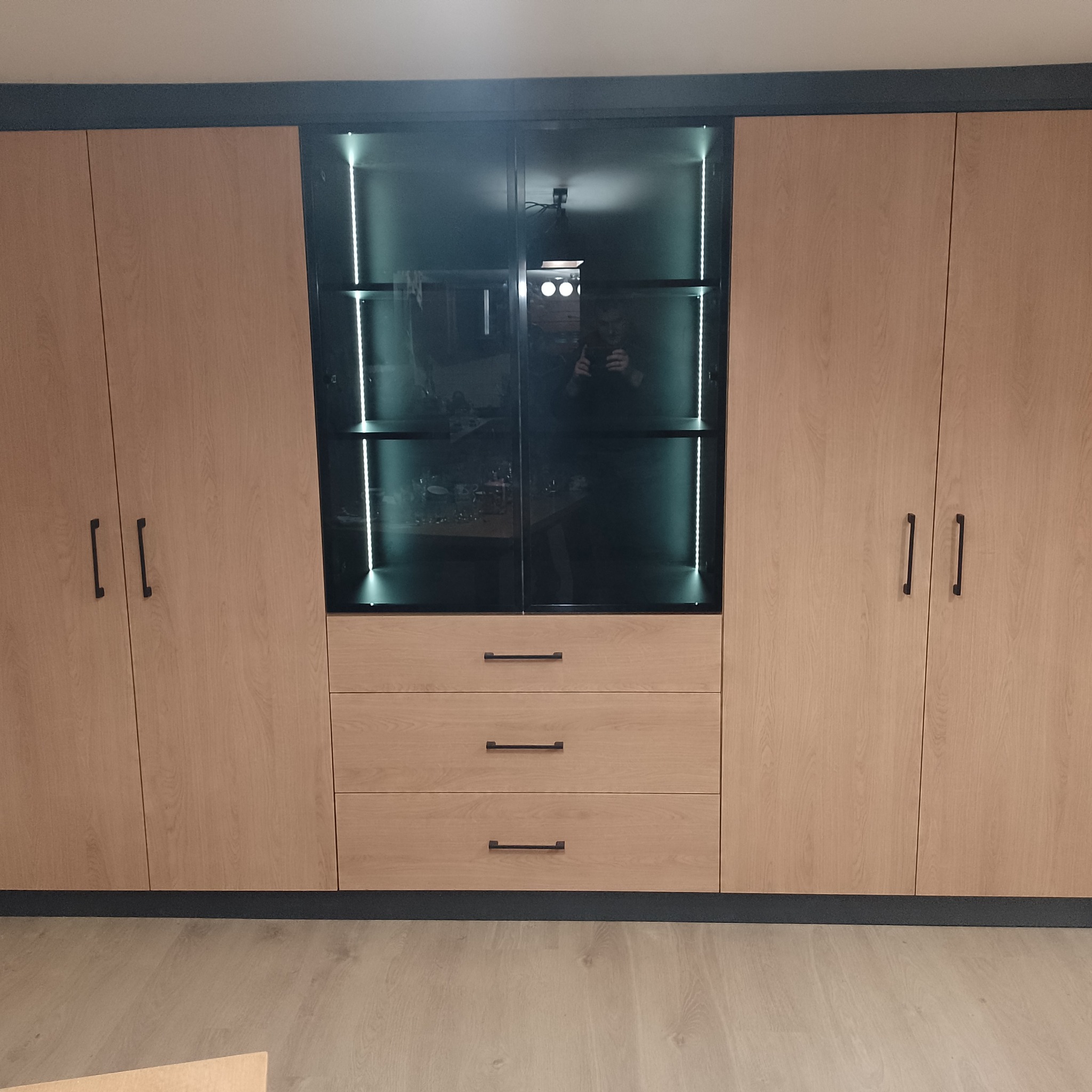 Jasna, drewniana szafa z trzema szufladami i przeszkloną witryną z oświetleniem LED wewnątrz, czarne, minimalistyczne uchwyty, widoczne odbicie fotografa w szkle.