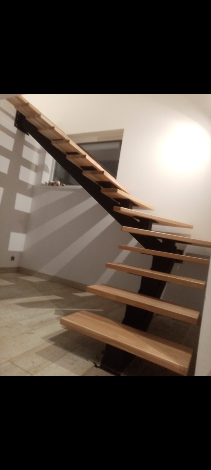 Drewniane schody z czarną metalową konstrukcją wspornikową, widoczne z dołu, z oknem w tle i marmurową posadzką.